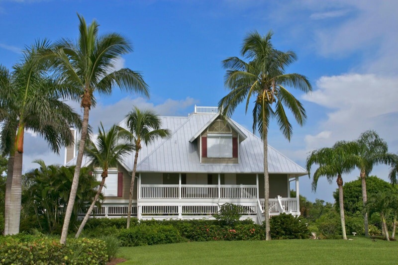 A tan Florida-style home with a silver-gray standing seam metal roof, a red-shuttered dormer, and a wraparound porch